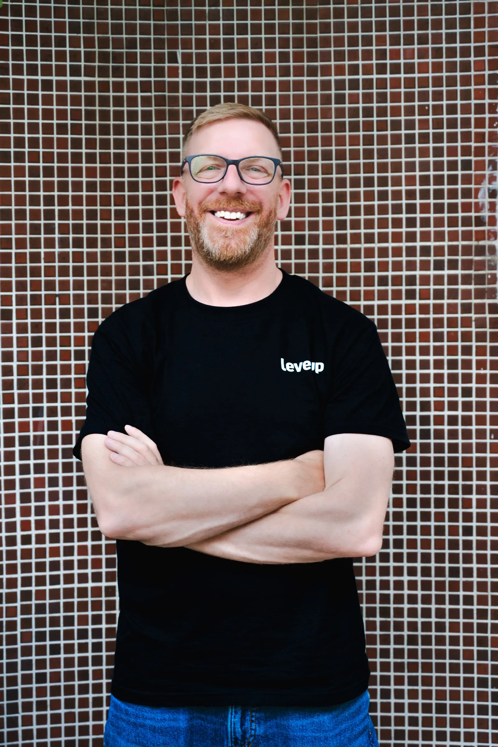Chris Winfield-Blum standing arms crossed in a black LevelUp T-shirt against a tiled wall